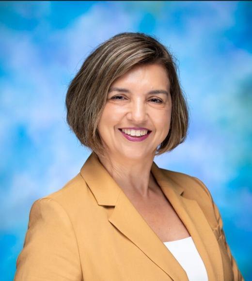 Headshot photograph of a smiling middle-aged woman in a mustard blazer, blue-green backdrop.